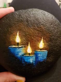 a person holding up a painted rock with three lit candles on it