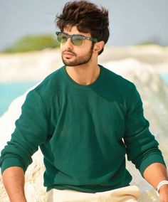 a man sitting on top of a rock next to the ocean wearing sunglasses and a green sweater