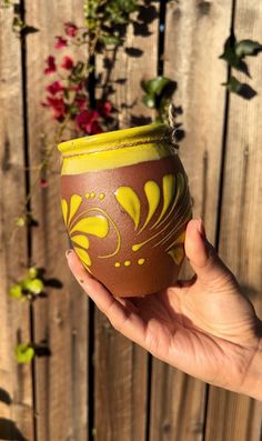 a hand is holding a yellow and brown vase in front of a fence with flowers