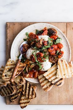 a white plate topped with toasted bread and salad next to crackers on a wooden cutting board