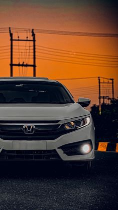 the front end of a white car parked in a parking lot at sunset with power lines behind it