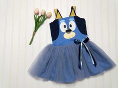 This kids Blue Heeler character dog dress is a charming and playful outfit perfect for little ones. The dress features a vibrant shade of blue, adding a pop of color to any occasion.  It has a character face and ears adorning the bodice.  The skirt has tulle over the cotton fabric adding a touch of whimsy and fun.  The back of the bodice has elastic encased for ease of dressing and will stretch for extended wear. Also available is little sister Orange Heeler. Please select Orange or Blue in the dropdown for your desired character. Sizes available are from 6 months to size 6. *Please allow two weeks from your date of order for your dress to ship.* Blue Disney Dress For Costume Party, Playful Fitted Cartoon Print Dress, Cute Blue Cartoon Print Dress, Cute Blue Dress With Cartoon Print, Blue Fitted Disney Dress, Playful Cotton Dresses With Character Print, Playful Cotton Dress With Character Print, Fitted Fun Blue Dress, Fun Fitted Blue Dress