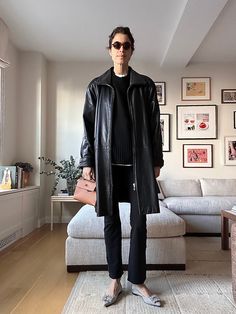 a woman standing in a living room wearing a black leather coat and silver shoes with her hands on her hips