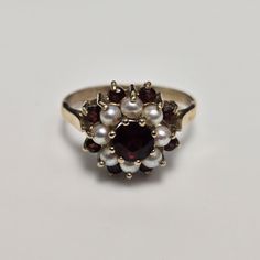 9ct Yellow Gold Garnet and Pearl Ring | UK Ring Size N | Elegant & Unique Elegantly crafted from 9ct yellow gold, this ring boasts a captivating combination of a vibrant garnet and lustrous pearls. The central garnet gemstone, measuring 4.5mm x 4.5mm, is surrounded by eight exquisite pearls, creating a timeless and sophisticated design that exudes charm and elegance.    Features:    Metal: 9ct Yellow Gold Gemstones: 1 Garnet & 8 Pearls    Center Gemstone Size: 4.5mm x 4.5mm    Hallmarks: Authent Garnet And Pearl, Photo Ring, Pearl Gemstone, Garnet Rings, Garnet Gemstone, Yellow Gold Ring, Sophisticated Design, Multi Stone Ring, Pearl Size