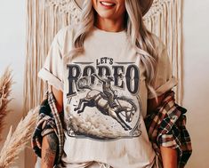 Channel the electrifying thrill of the rodeo with Lasso & Lace's "Let's Rodeo" t-shirt. This captivating design features a dynamic scene of a cowboy in full motion, expertly riding a bucking bronco, encapsulated within bold, vintage lettering that proudly declares, "Let's Rodeo". The artwork is skillfully printed on the top-tier Bella Canvas 3001 tee, renowned for its comfort and durability, making it a perfect fit for both active days and relaxed evenings. Available in three soft hues--White, N Crew Neck Letter Print Shirt For Rodeo, Bucking Bronco, Disco Cowgirl, Aesthetic Gift, Western Graphic Tees, Rodeo Shirts, Cowgirl Aesthetic, Cowgirl Shirts, Country Shirts