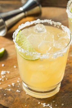 two margaritas sitting on top of a wooden cutting board next to lime wedges