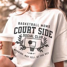 a woman holding a cup of coffee and wearing a t - shirt that says basketball moms court side social club