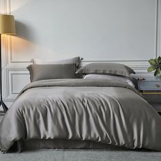 a bed with grey sheets and pillows next to a lamp on a nightstand in a white room