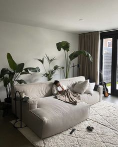 a person laying on a couch in a living room next to a large window with plants