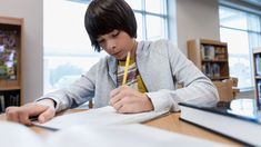 a young boy sitting at a desk writing on paper with a pencil in his mouth