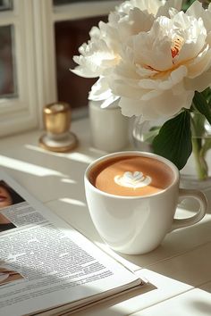 there is a cup of coffee on the table next to a magazine and some flowers