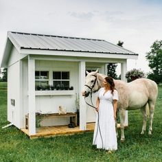 a woman in white dress standing next to a horse near a small shed with chicken coop