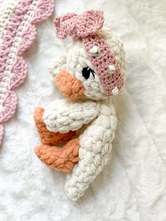 a crocheted stuffed duck laying on top of a white bed next to a pink and white blanket