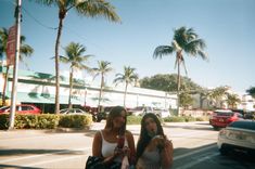 two women standing in front of a building with palm trees and cars on the street