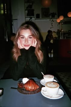 a woman sitting at a table in front of a cup of coffee and croissant