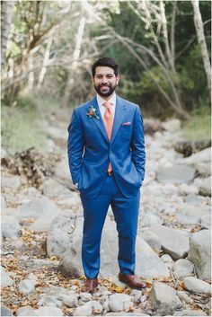 a man wearing a blue suit and orange tie standing on rocks in front of trees