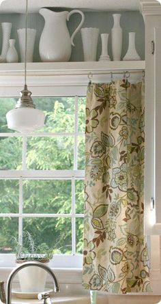 a kitchen window with curtains and dishes on the shelf above it, in front of a sink