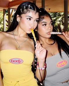 two young women standing next to each other holding food sticks in their mouths and looking at the camera