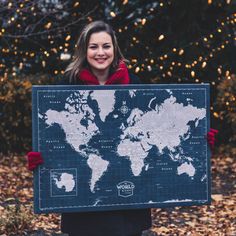 a woman holding up a world map in front of trees with lights behind her and leaves on the ground