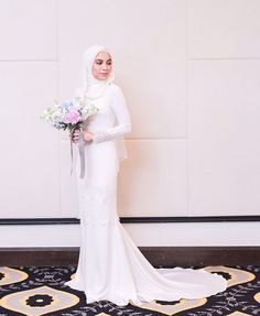 a woman in a white wedding dress holding a bouquet and standing next to a wall