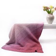 a pink and purple blanket sitting on top of a white table next to a vase with flowers