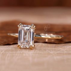 an emerald - cut diamond ring sits on a piece of wood, next to it's own wedding band