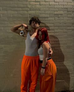 two people standing next to each other in front of a brick wall, one with red hair and the other wearing orange pants