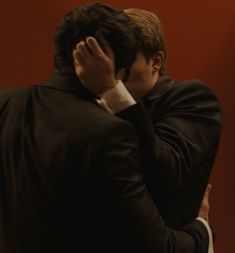 a man in a suit covers his face with his hands as he stands against a red wall