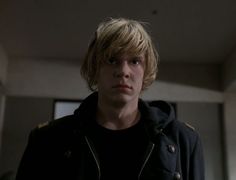 a young man with blonde hair wearing a black jacket and standing in front of two windows