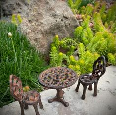 two miniature chairs and a table in front of some plants, rocks and flowers on the ground