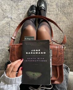 a person sitting on a couch holding a book in front of their feet and purse