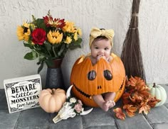 a baby in a pumpkin costume sitting next to some flowers and other halloween decorations on the ground