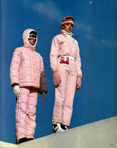 two people in pink snowsuits standing next to each other on a ramp with their feet up