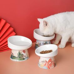 a white cat eating food out of two bowls on a table next to red paper fans