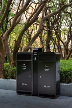 two black trash cans sitting next to each other on the side of a road near trees