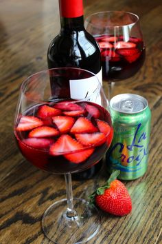 two wine glasses filled with red liquid and strawberries next to a bottle of soda