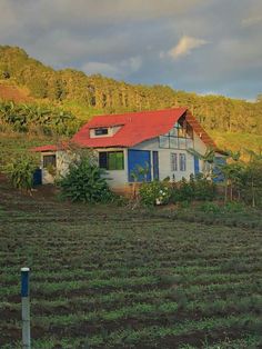 a house in the middle of a field with trees and hills behind it on a cloudy day