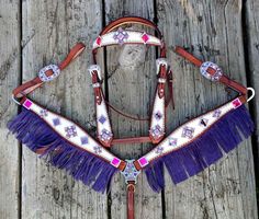 a purple and white horse bridle with fringes on the sides is sitting on a wooden surface