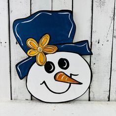 a snowman with a blue hat and yellow flower on it's head is standing in front of a white fence