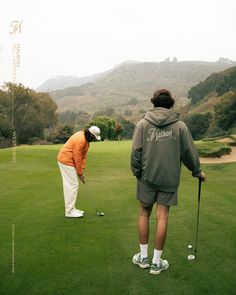 two men playing golf on a green field with mountains in the background and one man wearing an orange hoodie