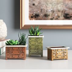 three small wooden drawers with plants in them on a table next to a framed painting