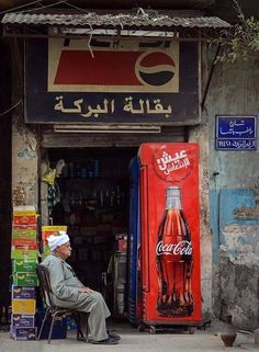 an old man sitting on a chair in front of a coca cola machine