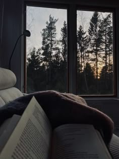 an open book sitting on top of a bed next to a window with trees outside