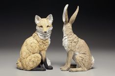 two figurines of foxes sitting next to each other on a gray surface with black background