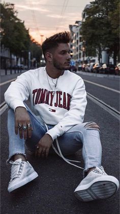 a young man sitting on the ground with his feet crossed wearing white sneakers and a tommy jean sweatshirt