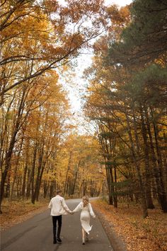 a man and woman holding hands while walking down a road surrounded by trees in the fall