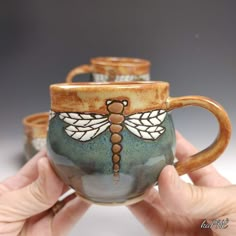 two handmade mugs with dragonfly designs on the handles and sides are held in front of each other