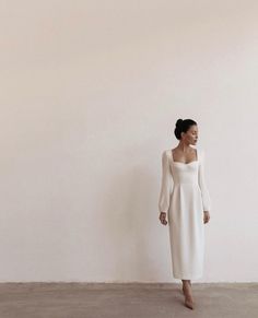 a woman standing in front of a white wall wearing a long sleeved dress and heels