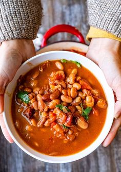 two hands holding a bowl of beans and meat soup with garnish on top