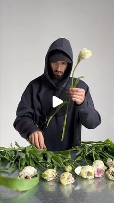 a man in a black hoodie is holding a flower and arranging it on a table